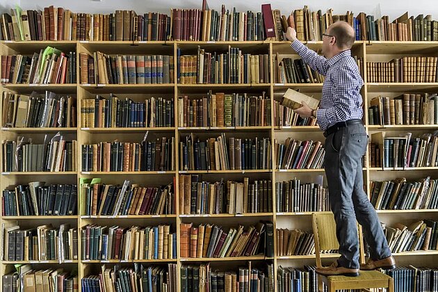 Per Anders Rudling står på en stege i ett bibliotek och håller på att ta en bok.