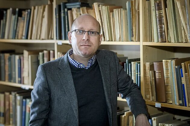 Per Anders Rudling i ett bibliotek. Hanstår rakt fram och tittar in i kameran.
